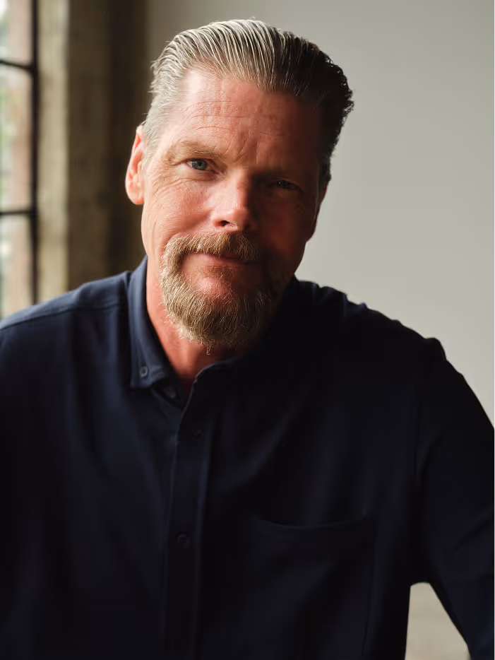 Middle-aged man with slicked-back hair and beard wearing a dark shirt near a window.