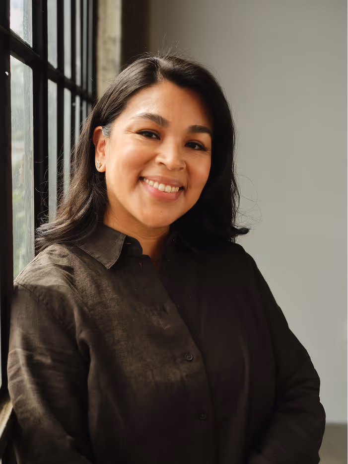 Smiling woman with dark hair wearing a dark brown shirt, standing by window with black frames.
