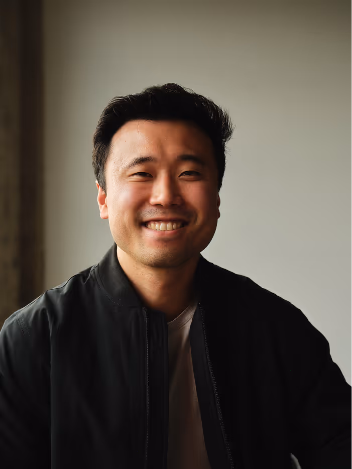 Smiling Asian man in a black jacket with a neutral gray background.