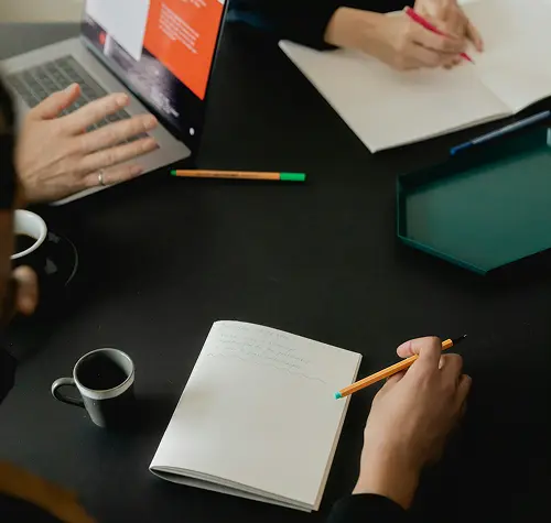 People working at a black table writing in notebooks with pencils, laptop, and coffee cups nearby.