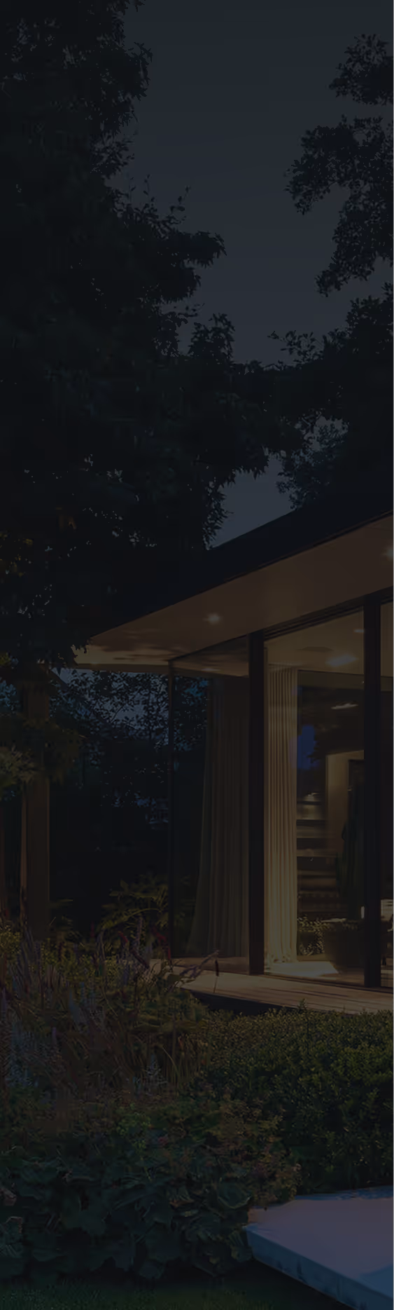 Modern house with large glass windows illuminated at night, surrounded by trees and garden.