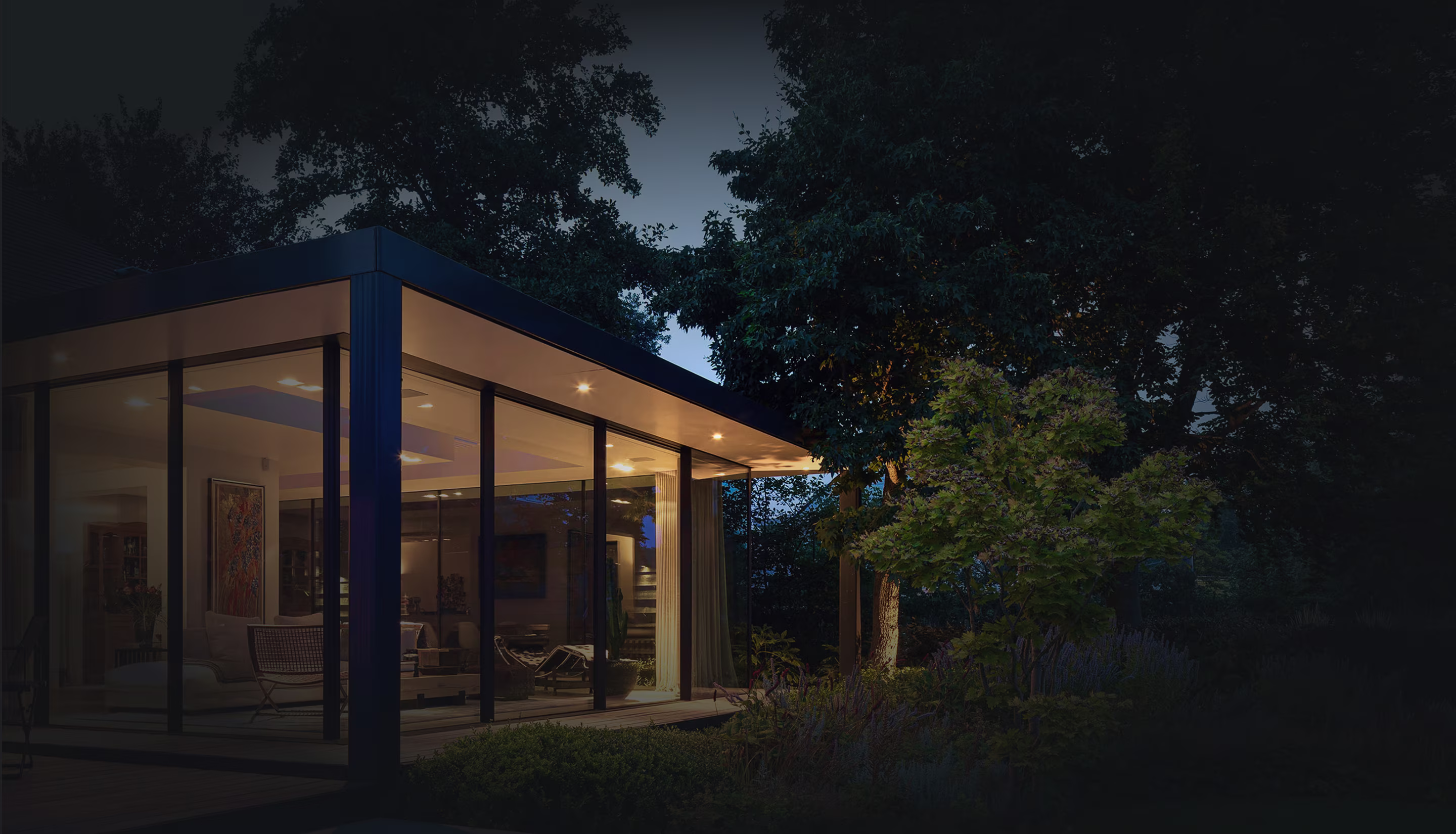 Modern glass-walled house lit warmly at dusk with surrounding trees and garden.