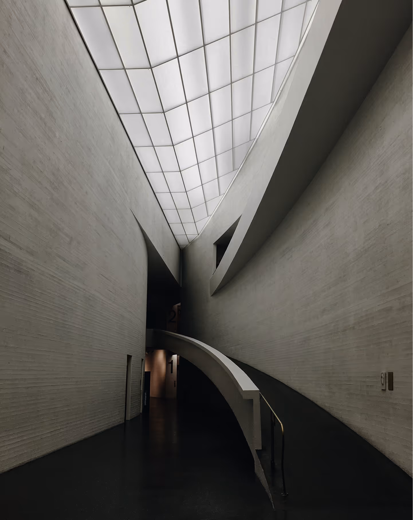 Interior view of a modern building corridor with high textured walls, a curved ramp, and a large skylight ceiling.